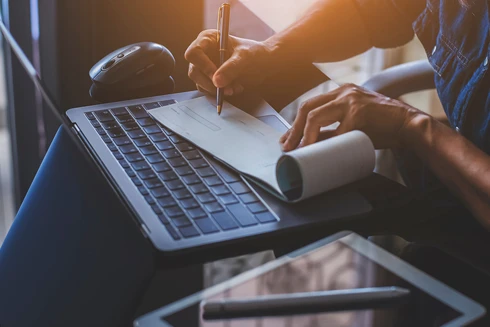 pormenor de mãos a assinar cheque, numa secretária com computador, tablet e caneta, com luz de pôr do sol