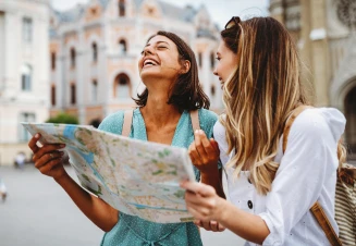 Duas mulheres jovens a sorrir e a segurar um mapa de papel enquanto exploram uma praça europeia com edifícios históricos ao fundo.