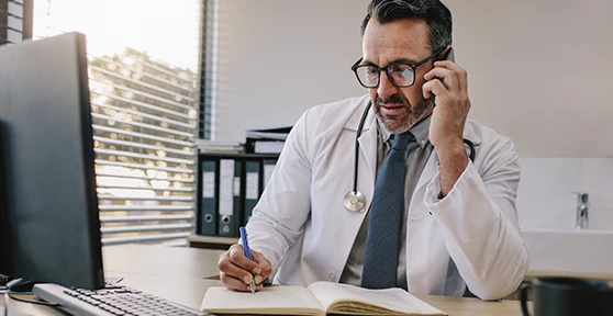 médico a atender paciente por telemóvel, a escrever num caderno e com computador em frente