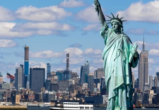 Vista panorâmica de Nova Iorque com a Estátua da Liberdade em primeiro plano à direita e os arranha-céus de Manhattan ao fundo sob um céu azul.