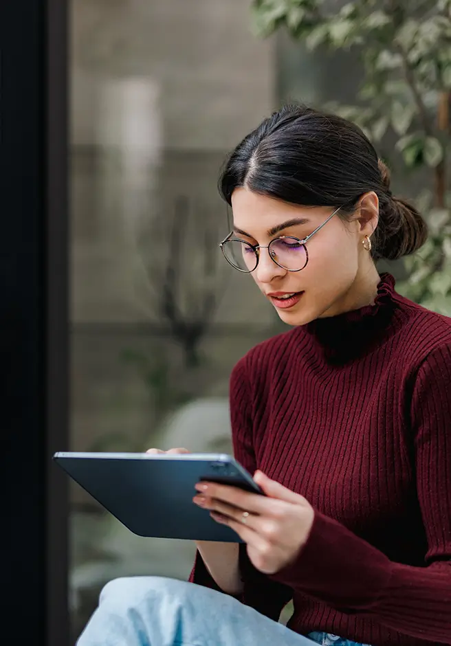 young woman sitting in front of a window with plants, using a tablet