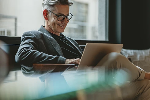 homem sorridente sentado num sofá à janela, com portátil ao colo