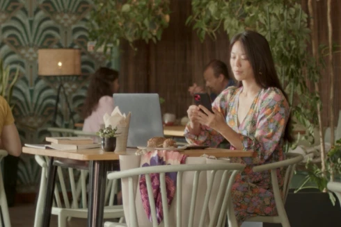 Rapariga na casa dos 30 anos com um vestido com flores a olhar para o seu telemóvel e computador portátil, numa mesa de um café