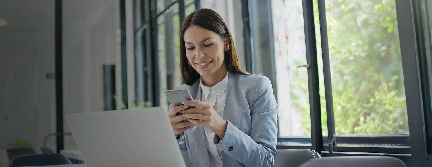 Mulher sorridente a usar o telemóvel num escritório com um portátil à sua frente