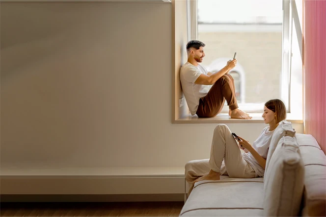 Casal jovem a consultar telemóvel. Ele sentado na janela e ela no sofá