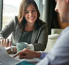 Homem e mulher a conversar, sentados a uma mesa com papéis, um computador e uma caneca de café