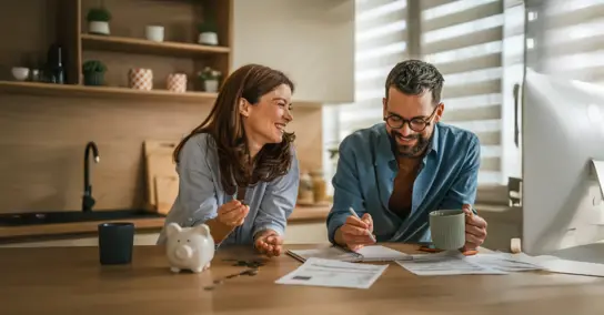 Casal a sorrir na cozinha a analisar documentos e um computador portátil
