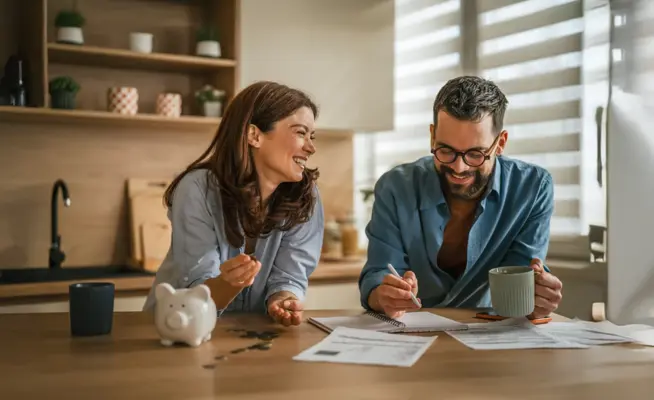 Casal a sorrir na cozinha a analisar documentos e um computador portátil