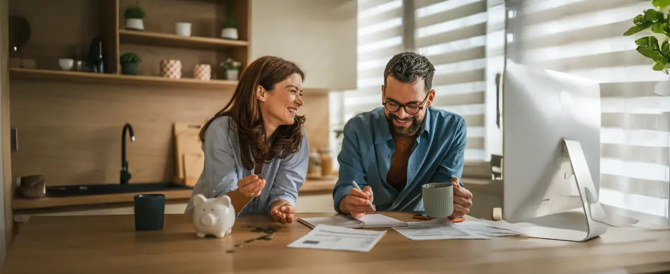 Casal a sorrir na cozinha a analisar documentos e um computador portátil