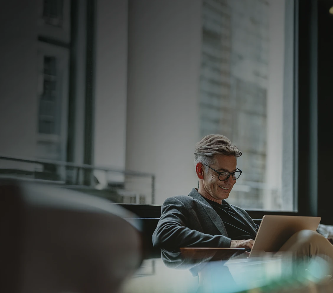 smiling man sitting on a sofa by the window with a laptop on his lap