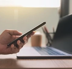 Mão a segurar um telemóvel em frente a um computador portátil