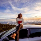 Mulher sentada no tejadilho de um carro observa o pôr do sol sobre as nuvens