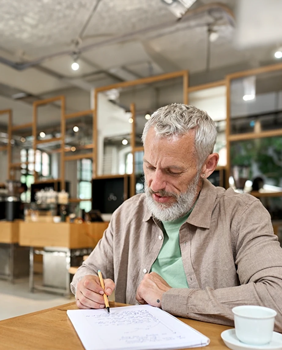 Senhor sentado à mesa, a tomar notas num caderno e com chávena em cima da mesa