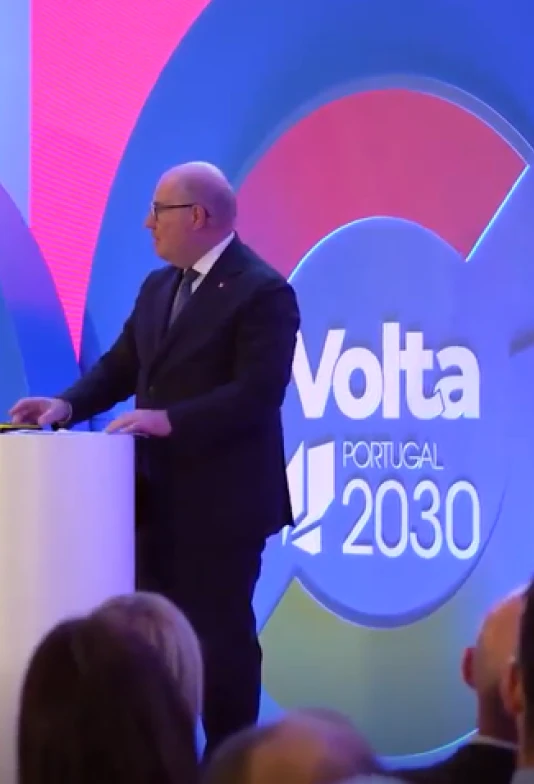 Homem, de óculos, fato e gravata, a dar uma palestra à frente de um cenário com o logo "Volta Portugal 2030"