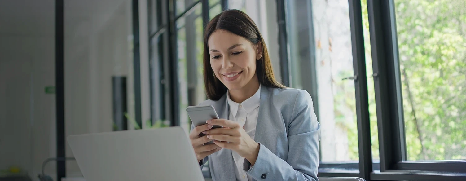 Smiling businesswoman using her mobile phone in an office with a laptop in front of her