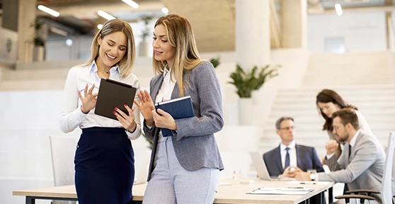 2 mulheres numa sala de reuniões a conversar, com 3 pessoas ao fundo