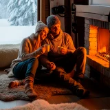 Casal sentado junto à lareira numa cabana de inverno, com paisagem nevada pela janela