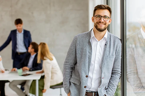homem sorridente encostado a janela de vidro, numa sala de reuniões com 4 pessoas atrás