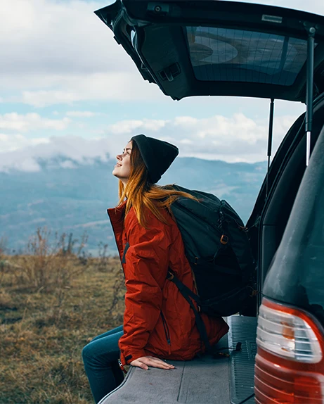 mulher sentada na bagageira do carro, a olhar para paisagem com montanhas