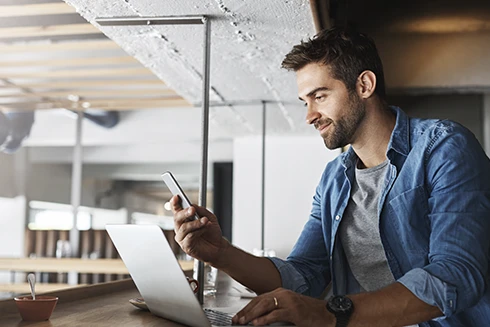Jovem rapaz sentado em frente ao computador, com o telemóvel na mão e a sorrir