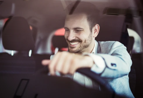 Homem de camisa branca e blazer cinza ao volante de um carro a sorrir para o tablier