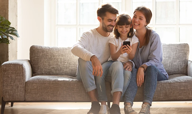 Jovem família sentada no sofá: pai, mãe e filha, sorrindo e olhando para telemóvel