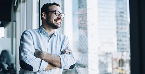 Man with glasses smiling with arms crossed by a window