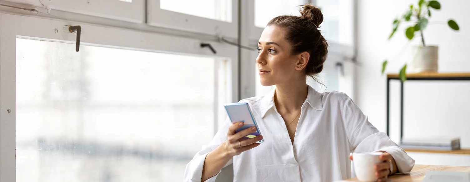 lady looking out the window, with her phone in one hand and holding a cup with the other hand