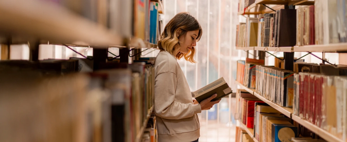 Mulher de pé entre estantes de uma biblioteca a ler um livro