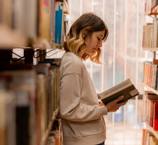 Mulher de pé entre estantes de uma biblioteca a ler um livro