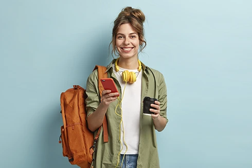 rapariga sorridente, com mochila, telemóvel na mão, e headphones à volta do pescoço