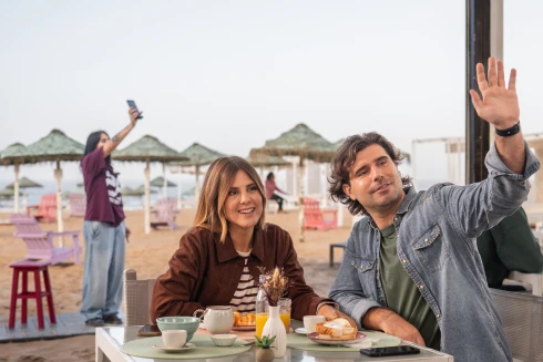 Um homem e uma mulher sentados num bar de praia, e o homem a acenar a alguém