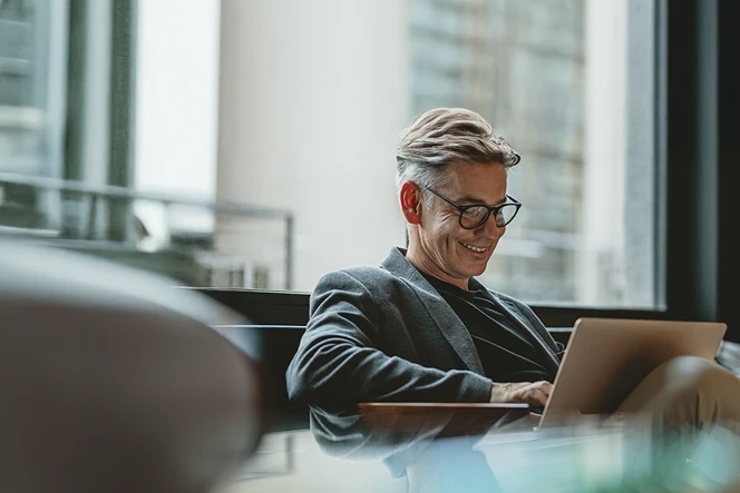 smiling man sitting on a sofa by the window with a laptop on his lap