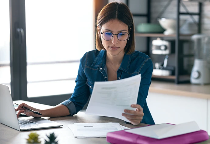 senhora sentada em frente a computador, com papéis na mão