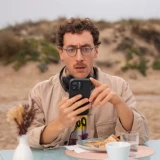 Jovem rapaz, sentado em mesa de esplanada em praia, com ar de espanto a olhar para o telemóvel