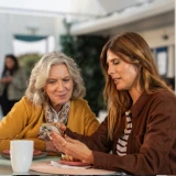 Duas mulheres, sentadas numa mesa, a olharem para o telémovel