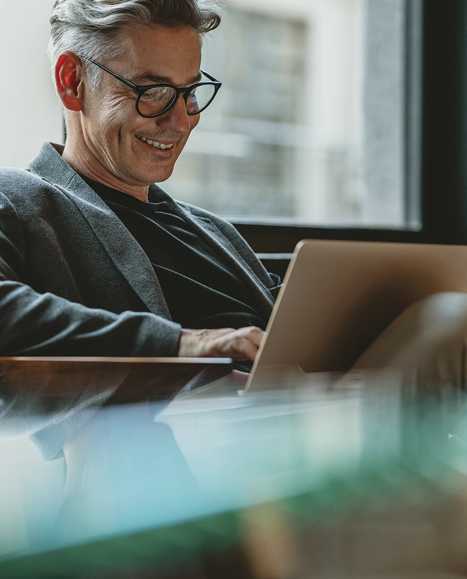 homem sorridente sentado num sofá à janela, com portátil ao colo