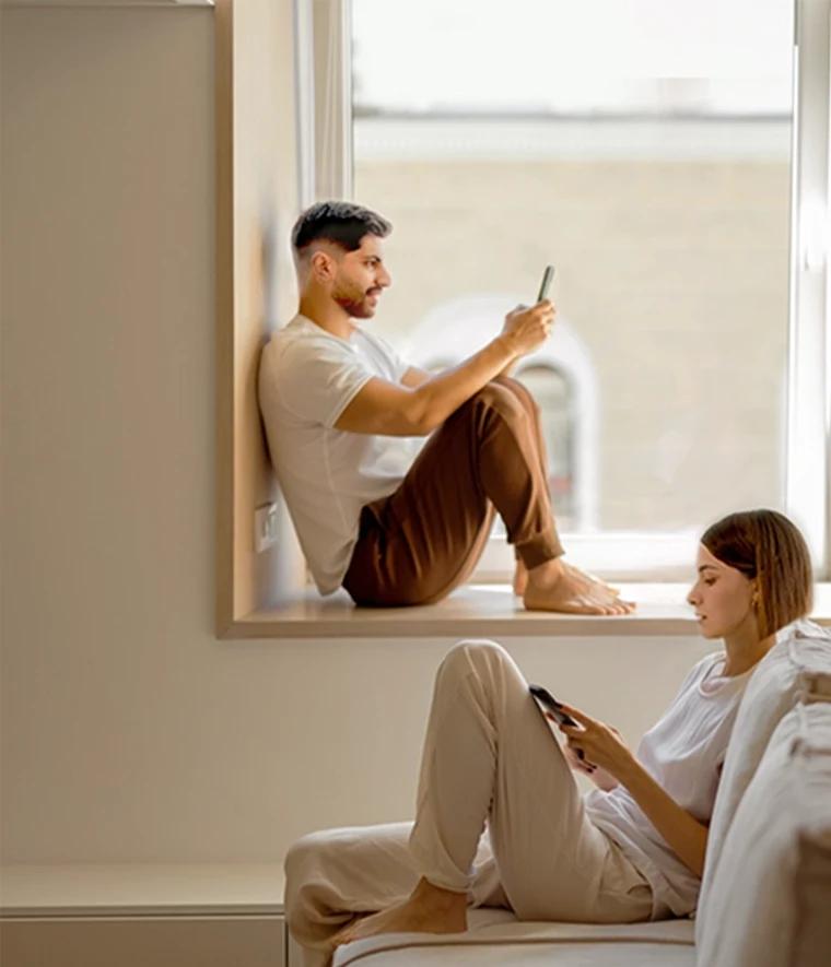 young couple checking their phones. He's sitting by the window and she's on the sofa