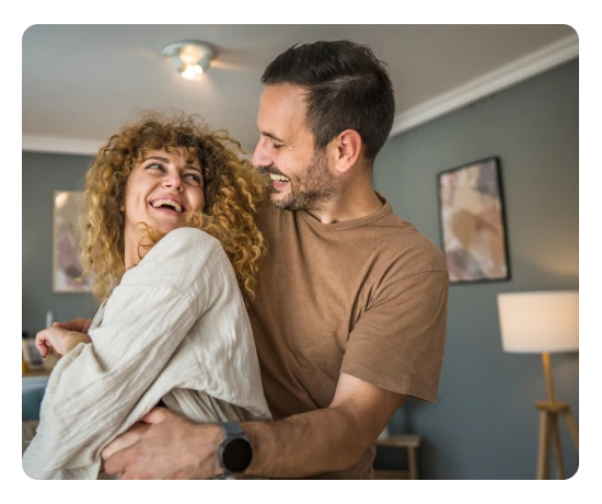 Casal a rir e a abraçar-se descontraidamente no interior de uma sala de estar moderna e acolhedora.