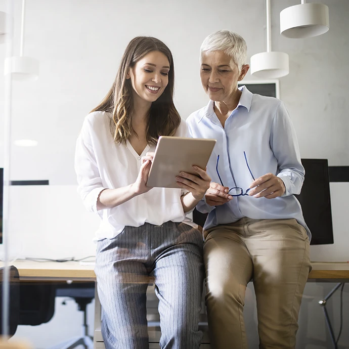 2 mulheres encostadas a uma secretária, a conversar, e a ver um tablet