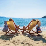 casal sentado em cadeiras amarelas numa praia de mar azul, numa ilha com montanha verde
