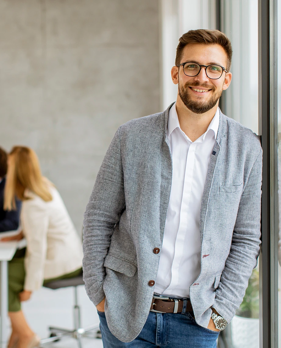 homem sorridente encostado a janela de vidro, numa sala de reuniões com 4 pessoas atrás