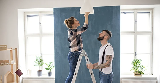 couple, she on a stepladder changing a light bulb in a lamp, he holding the stepladder