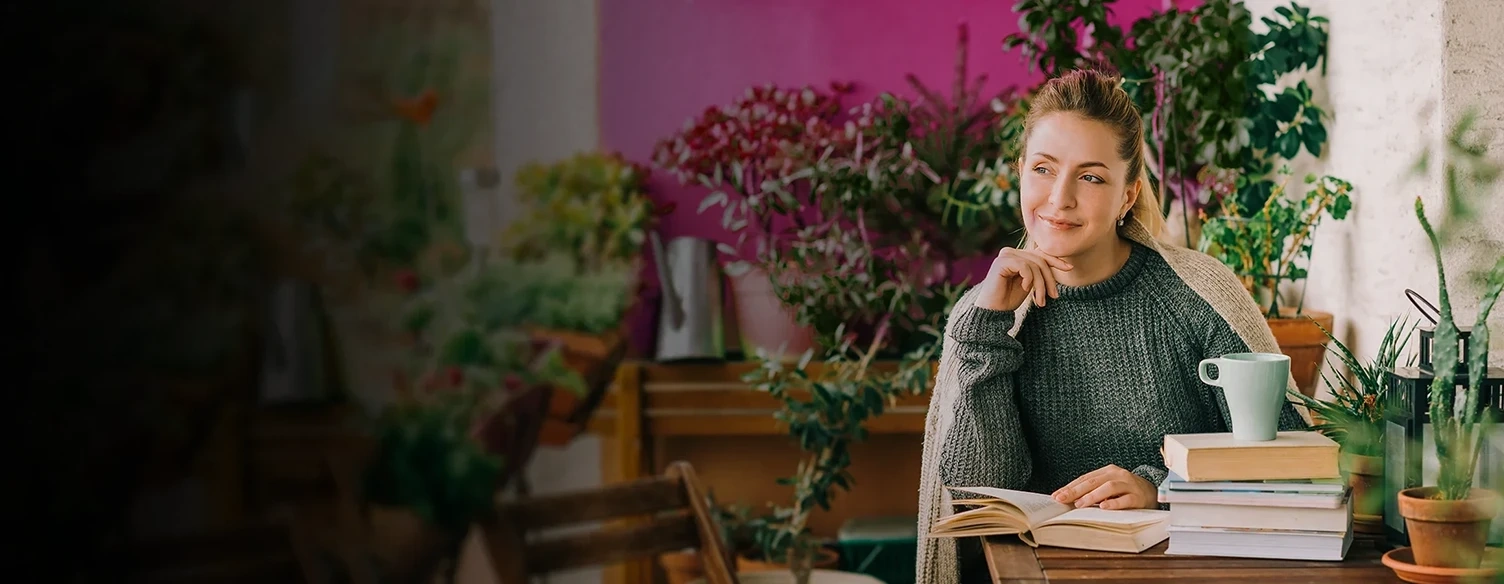 Relaxed woman sitting at a table with books and plants, looking thoughtfully to the side