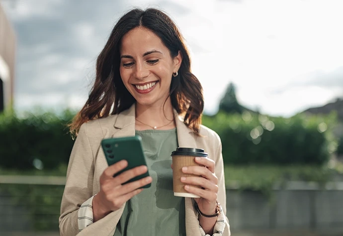 Mulher sorridente a usar o telemóvel enquanto segura num copo de café