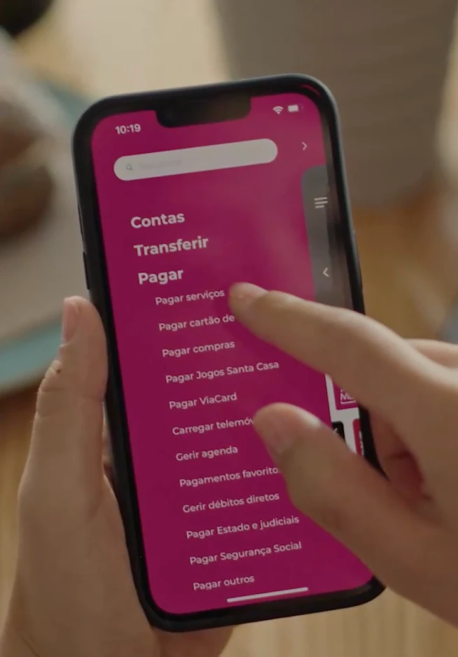 Mão a agarrar um telemóvel que no seu ecrã apresenta o menu da App Millennium, com destaque para a secção "Pagar"