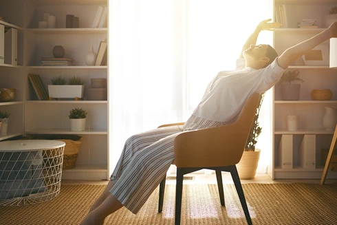 mulher sentada numa poltrona na sala, a espreguiçar-se, com luz do sol a entrar pela grande janela