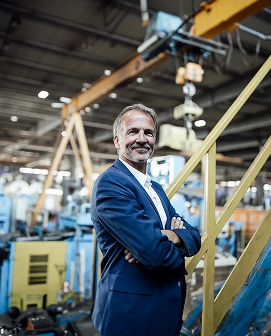 homem vestido de fato azul, com braços cruzados e sorridente, numa fábrica