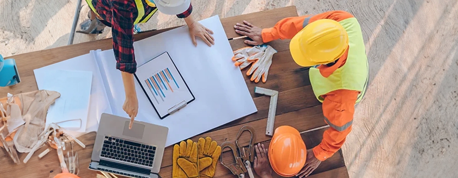 plano superior de trabalhadores da construção civil à volta de mesa com portátil, plantas, luvas e capacetes